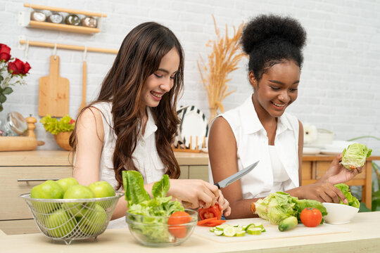 Black and white teenage friends happily and joyfully together to making a vegetable salad in a white kitchen. They enjoy healthy food.