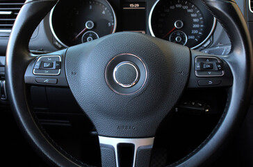 Steering wheel Close up. Modern black leather car steering wheel with multifunction control buttons and chrome detailing, shown in close-up with dashboard gauges and textured surface clearly visible.