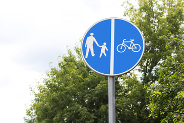 Shared pedestrian and bicycle path sign. Blue circular traffic sign with symbols. Urban infrastructure for walking and cycling. Safety road indication with green trees and cloudy sky in background.