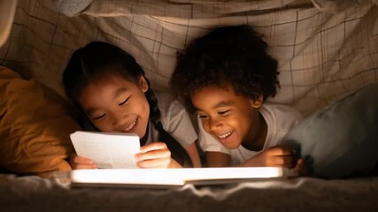 Happy children reading a book together under cozy blanket fort at night. - Powered by Adobe