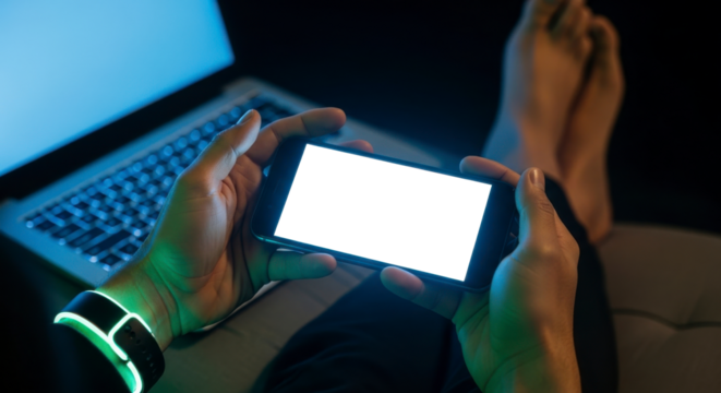 Human hands holding a smartphone with a glowing transparent screen displaying a laptop in the background representing modern technology and digital interaction in a dimly lit room