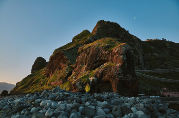 Person stands on a vast rocky beach gazing at towering green mountains and cliffs under a clear evening sky with a crescent moon, capturing the majesty of a rugged coastal landscape.