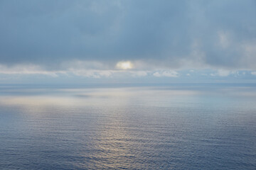 Sun peeking through a cloudy sky, casting a serene reflection on the vast, tranquil ocean surface at the horizon.