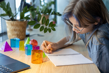 Teen girl studying 3D geometric shapes during math class, drawing figures in notebook and exploring spatial concepts through hands-on learning and interactive school project.