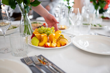 Elegant table setting with glassware and cutlery, as a hand places a plate of fresh sliced fruits including oranges, grapes, and apples, ready for a festive meal.
