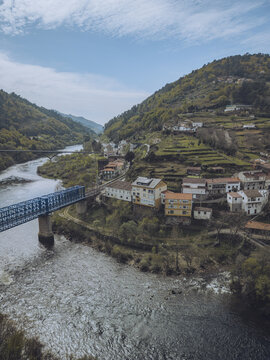 Aerial view of the blue bridge spans the textured river, connecting the quaint village nestled amidst the verdant hills, Os Peares, Ourense, Spain.