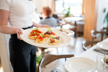 Waiter serving bruschetta with tomatoes, greens, and meat on a plate in a modern restaurant, showcasing professional hospitality and gourmet dining atmosphere.