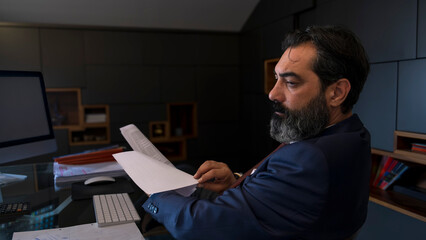 A middle-aged, dark-haired businessman in a suit is working seriously at his desk in the office,...