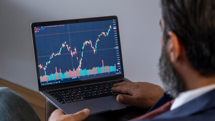 A middle-aged, dark-haired businessman in a suit is sitting on a leather couch in his office, monitoring graphics and working on his laptop