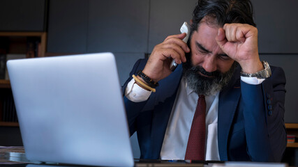 Middle-aged dark-haired businessman in a suit having a stressful business conversation on the phone while working with his computer at his desk in the office