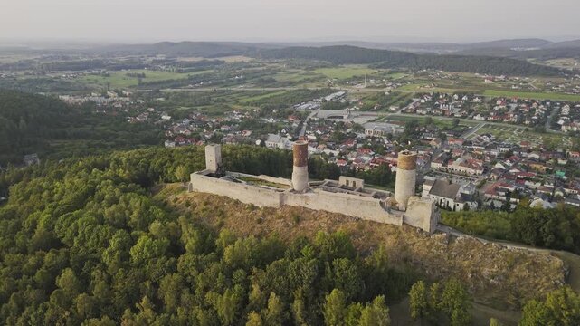 An impressive aerial view of Checiny Castle in Poland during summer. The medieval stone structure stands proudly on a hill, surrounded by lush greenery and clear skies.