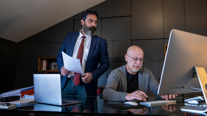 Two middle-aged businessman in a suit is in an office environment, working tensely with his computer and documents in front of him while holding a meeting