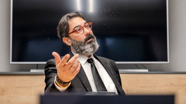 A middle-aged, dark-haired, bearded businessman in a suit is sitting at a conference table in his office, looking at his employees with a tense expression during a meeting
