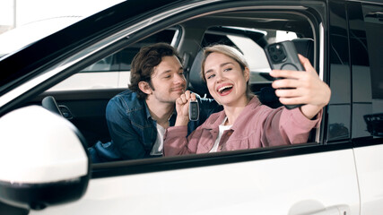 Fototapeta premium Young couple happily taking a selfie inside a new car at a dealership, showcasing excitement and joy of purchasing a vehicle