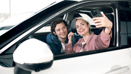 Fototapeta premium Young couple happily taking a selfie inside a new car at a dealership, showcasing excitement and joy of buying a vehicle together