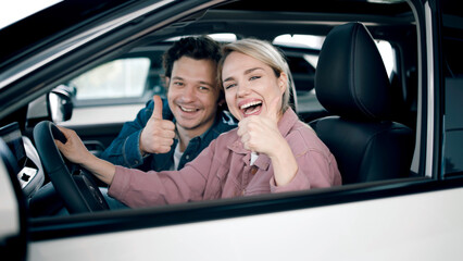 Happy couple inside car at dealership, showing thumbs up, celebrating their new purchase, enjoying the excitement of buying a vehicle