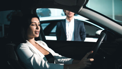 Woman in stylish outfit is seated in a car at a dealership, engaging with a male salesperson,...