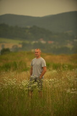 A man stands in a field of tall grass, holding a microphone as if he is about to speak or sing. The background features rolling hills and a cloudy sky.