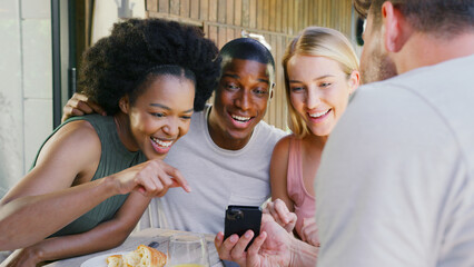 Group Of Smiling Multi-Cultural Friends Eating Breakfast Outdoors At Home Looking At Mobile Phone