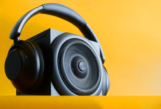 Black wooden hi-end acoustic speaker with metal grille paired with matching headphones on a bright yellow background conveying modern audio design and high-fidelity listening setups. Photo