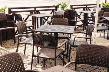 Empty outdoor café terrace with tables and chairs. Sunny day at restaurant patio. Urban dining space ready for guests. Brown plastic furniture on cobblestone ground. Relaxed atmosphere in daylight.
