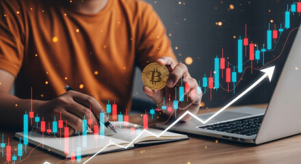 Person holding a Bitcoin cryptocurrency coin while writing in a notebook with a digital stock market graph overlay symbolizing investment and financial growth