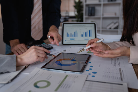 Business colleagues are pointing at charts displayed on a tablet and printed documents, collaborating on investment strategies and analyzing market trends during a productive office meeting