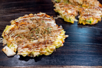 Traditional Japanese savory pancake okonomiyaki grilled on hot metal plate, finished with layered sauces and moving bonito flakes in the foreground