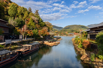 Fototapeta premium Wooden boats line the calm river in Arashiyama, Kyoto, Japan, with autumn trees on both banks and forested mountains in background under clear sky in early light