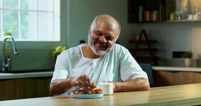 Senior Indian man eating pakoda or pakora with tea on table, Asian foodie old man expressing immense happiness while enjoying tasty onion fritters in modern kitchen setting, joyful retired person