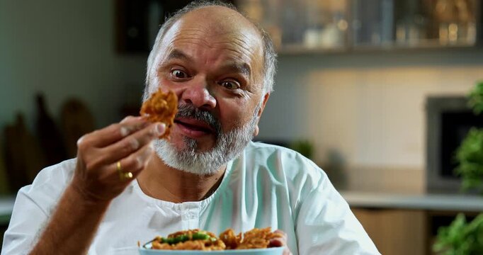 Senior Indian man eating pakoda or pakora with tea on table, Asian foodie old man expressing immense happiness while enjoying tasty onion fritters in modern kitchen setting, joyful retired person