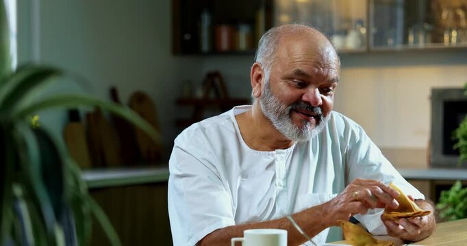 Senior Indian man eating samosa with tea at home kitchen, Asian foodie old man expressing immense happiness and joy while enjoying delicious Indian snack in modern domestic kitchen interior