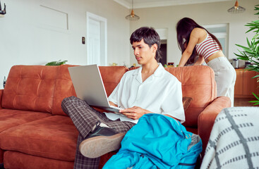 Young latin American digital nomad using laptop while sitting on sofa with backpack next to him, as woman standing in background