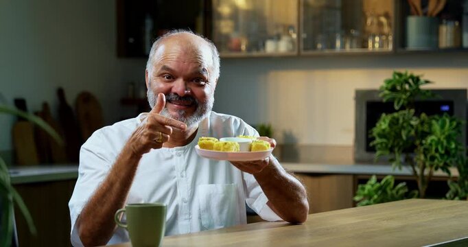 Senior Indian man eating khaman dhokla with joyful expression, Asian foodie old man enjoying tasty Gujarati snack while sitting at table in modern kitchen, expressing immense happiness and delight