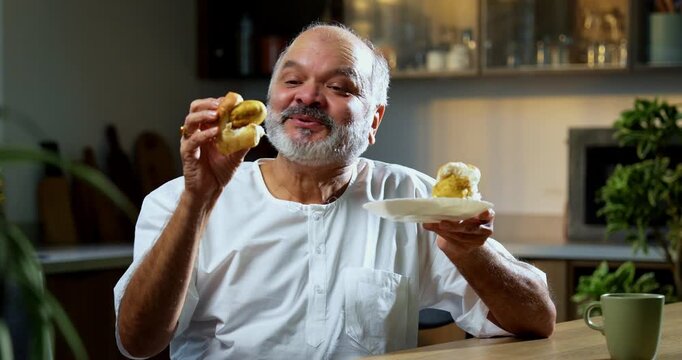 Senior Indian man eating vada pav with tea at home table, Asian foodie old man expressing immense joy and satisfaction while enjoying spicy Mumbai street food snack in modern Indian kitchen setup