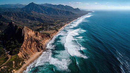 An aerial view captures dramatic cliffs along a sunlit coastline, with waves crashing against the shore and distant mountains rising in the background.