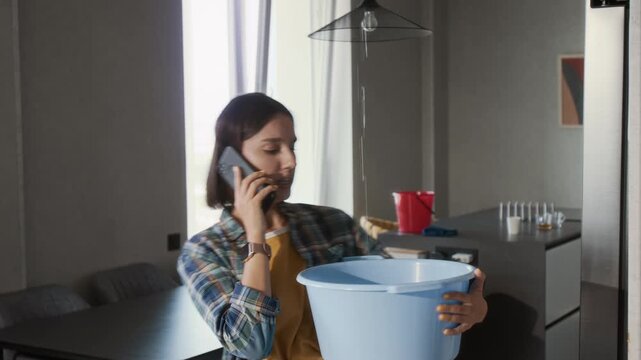Medium shot of young woman calling plumber by phone and holding plastic bucket catching water leaking from ceiling, copy space