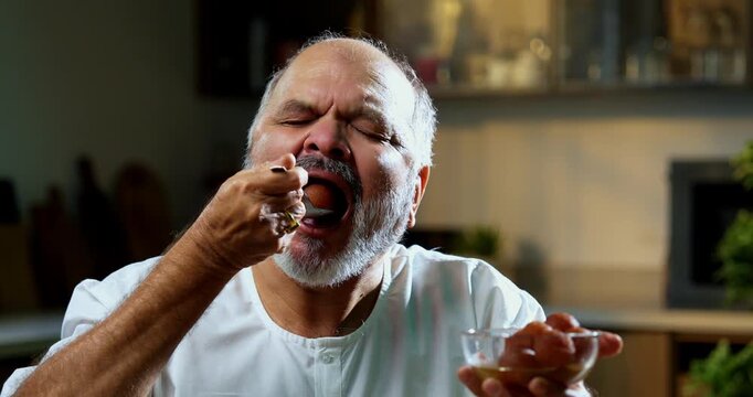 Senior Indian man eating sweet gulab jamun secretly at home, Asian foodie old man expressing immense joy, delight while enjoying delicious Indian dessert at table in bright modern kitchen interior