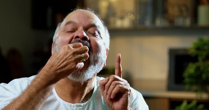 Senior Indian man eating sweet gulab jamun secretly at home, Asian foodie old man expressing immense joy, delight while enjoying delicious Indian dessert at table in bright modern kitchen interior