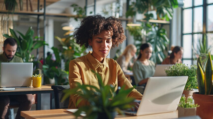 Young black woman working at a laptop in a modern, plant-filled office, surrounded by colleagues.