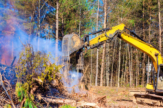 Excavator removes fallen trees, brush in forest as smoke billows from nearby fire during deforester day.
