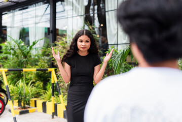 Curly-haired Asian woman in black raising hands in confusion in front of a man. Outdoor scene in...