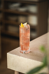 Pink cocktail with ice and lemon peel in tall glass on bar counter