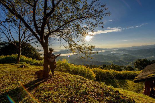 Mature man in traditional Thai clothing leaning against a tree in mountains playing the guitar, Chiang Rai, Thailand