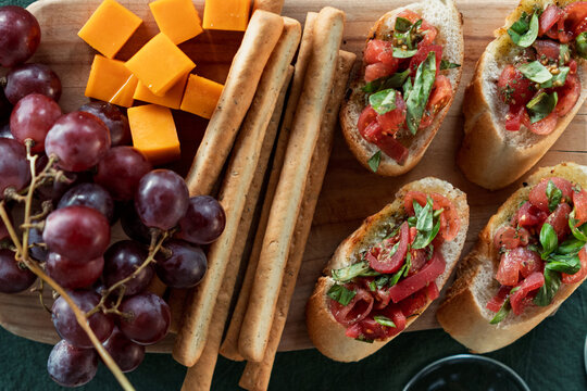 Overhead view of tomato bruschetta, cheese, grapes and breadsticks on a wooden chopping board