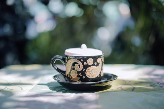 An elegant black and gold traditional batik cup and saucer, with a white lid, rests on a table. The intricate pattern stands out against a soft, sunlit green background.