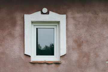 A small window on the wall of an old house, decorated with a white frame. The reflection of trees in the glass adds depth and mystery to the image. This is a symbol of coziness and tranquility, hidden