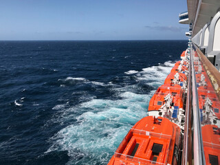 Orange lifeboats lined along the side of a cruise ship, viewed from a balcony with the deep blue ocean stretching out beyond, capturing both safety and serenity at sea.