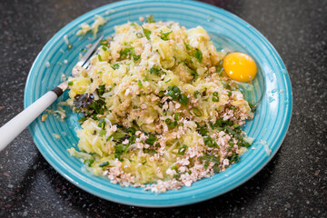 Hand mixing grated zucchini, oats, herbs and egg on blue plate preparing healthy dish. Zucchini pancakes