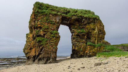 bering island. Natural rock formation with a large arch, surrounded by lush greenery and sandy beach, creating a stunning coastal landscape with dramatic textures and serene atmosphere